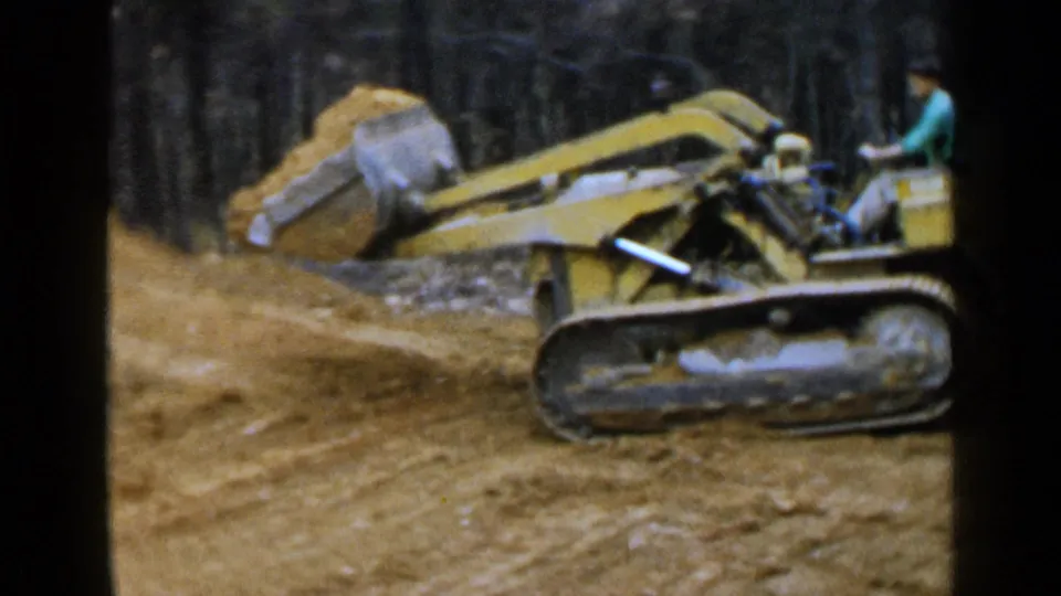 Side view of the yellow tracked loader carrying earth, with the operator seated at the controls in Birmingham, 1957.