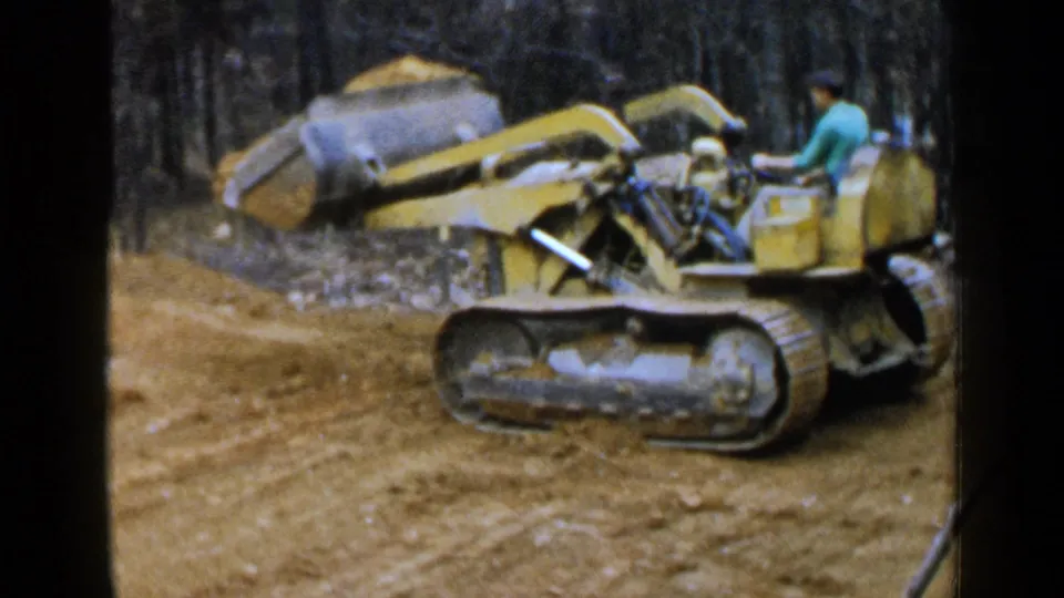 A man operates a yellow tracked loader with its bucket lifted at a muddy wooded Birmingham site in 1957.