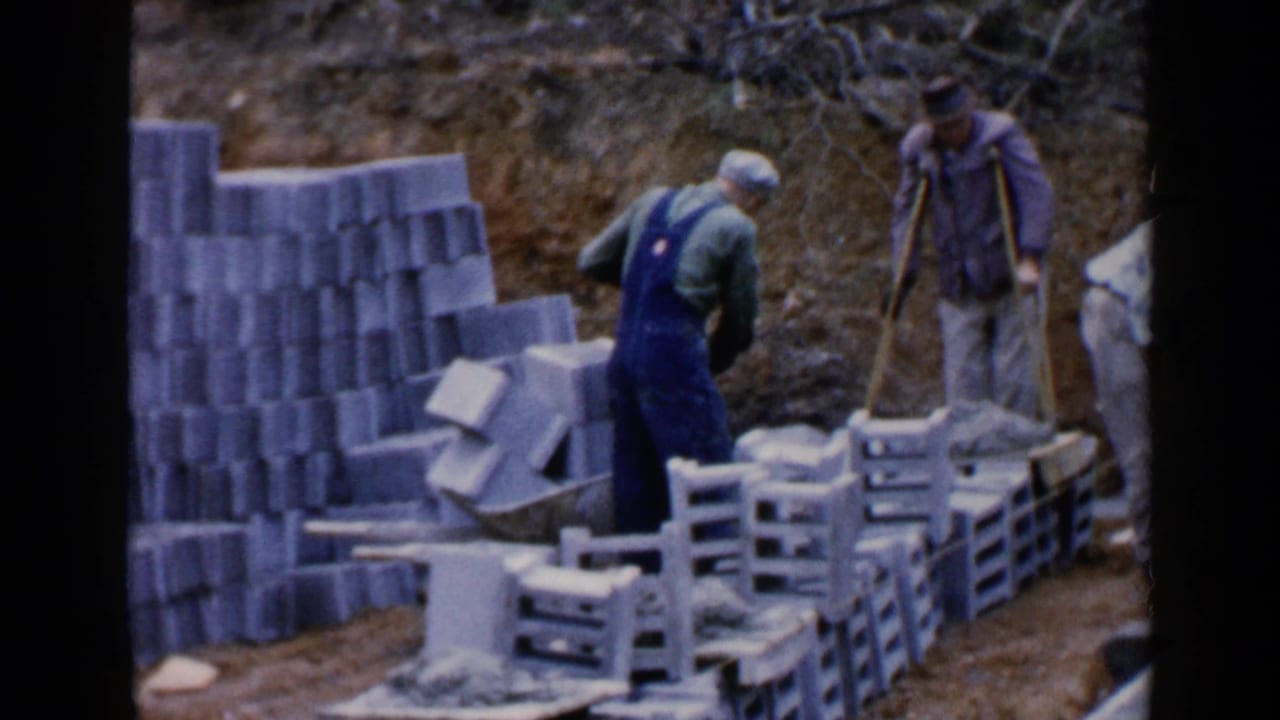 Workers stand near stacked concrete blocks and a wheelbarrow at a muddy construction site.