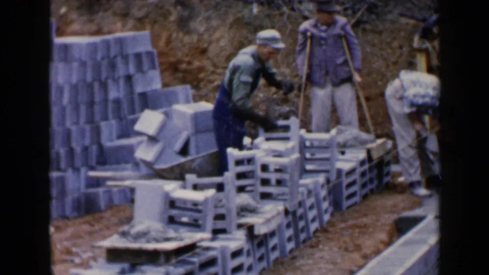 Adult workers in caps and overalls stand among concrete blocks and pallets at a Birmingham foundation site in 1957.