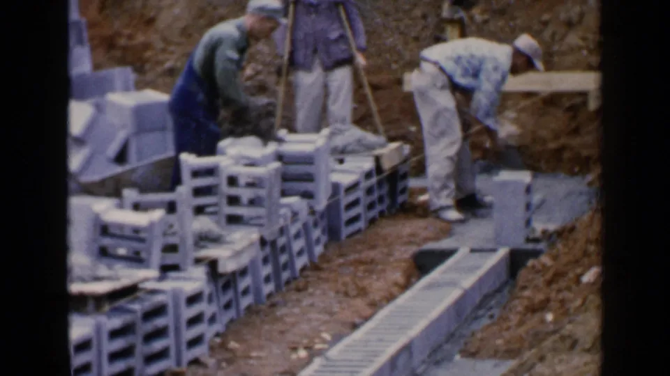 1957 Birmingham home-movie frame shows workers placing or preparing concrete blocks beside a narrow foundation trench.