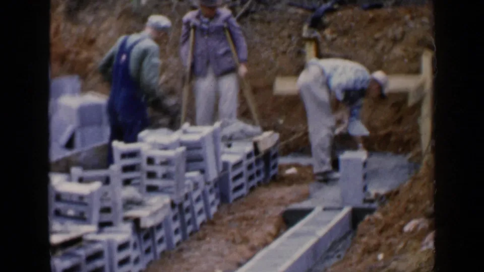 Adult men work with concrete blocks beside pallets and a trench in 1957 Birmingham construction footage.