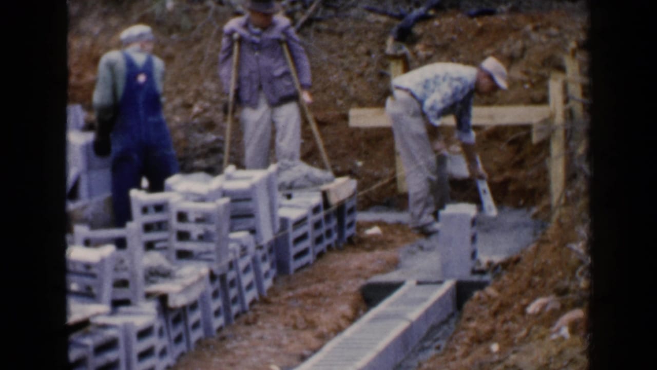 Workers place concrete blocks and cement in a muddy construction trench.