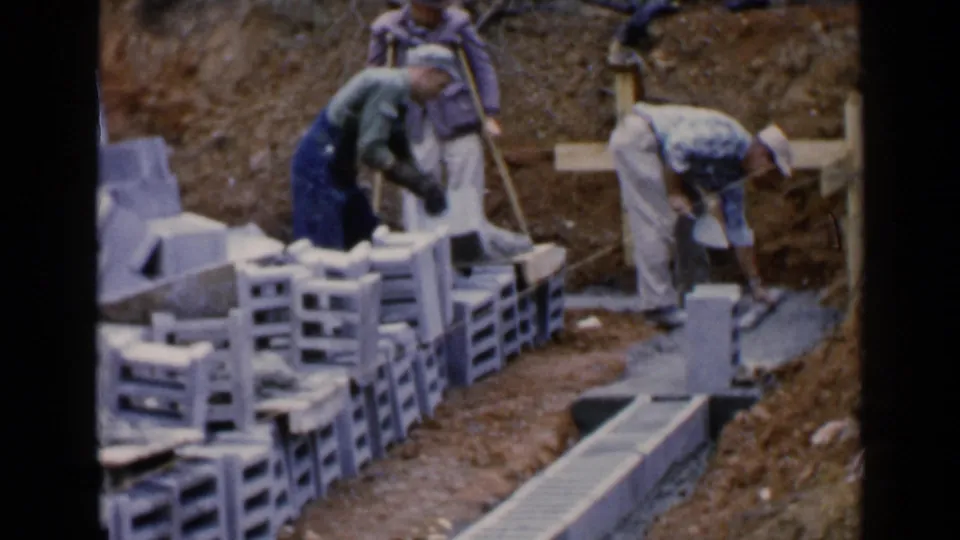 Workers in 1957 Birmingham handle concrete blocks near a trench as a man with crutches stands beside the work area.