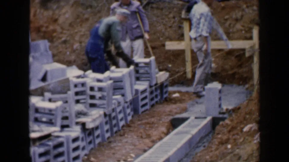 At a Birmingham construction site in 1957, a worker in overalls handles blocks while others stand near cement and wooden bracing.