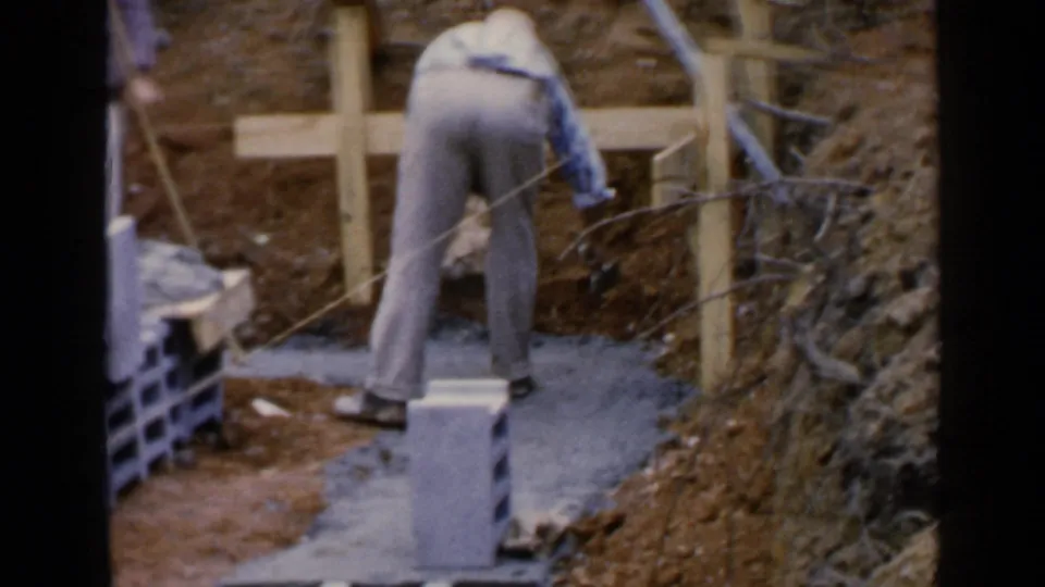A worker bends over cement beside concrete blocks and timber bracing at the 1957 Birmingham construction site.