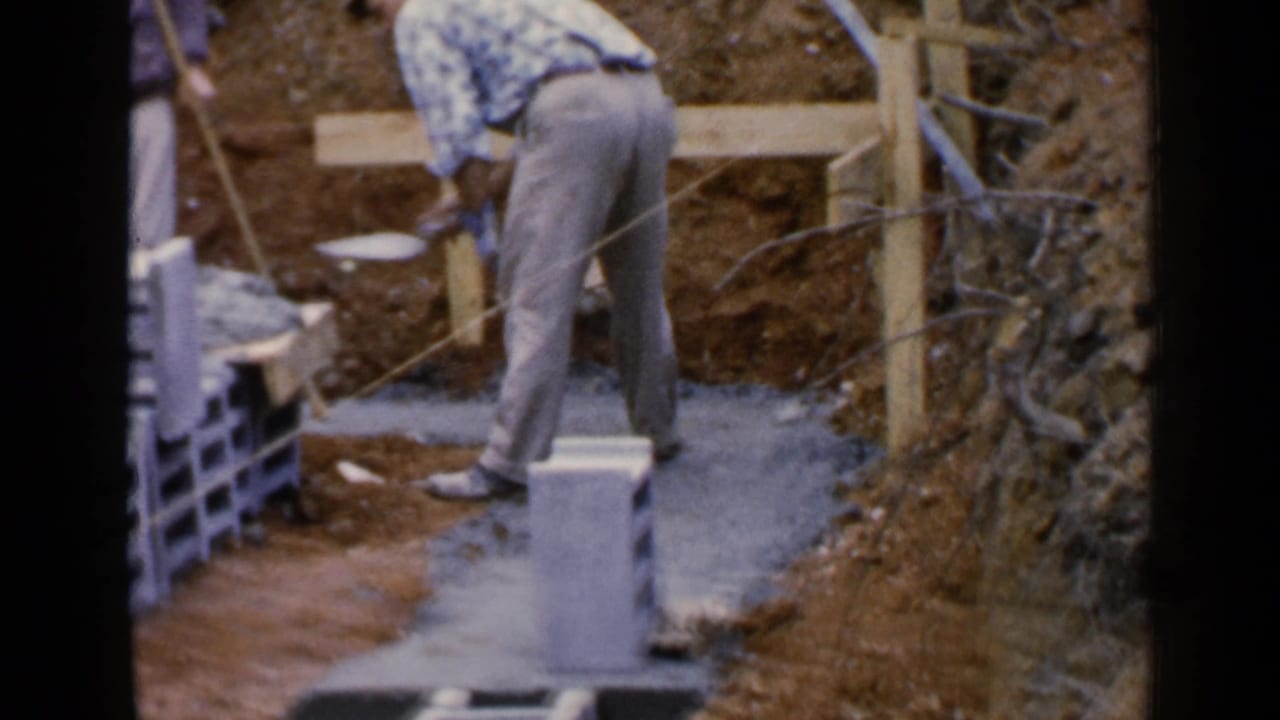 Man bends over wet cement near stacked concrete blocks and wooden formwork.
