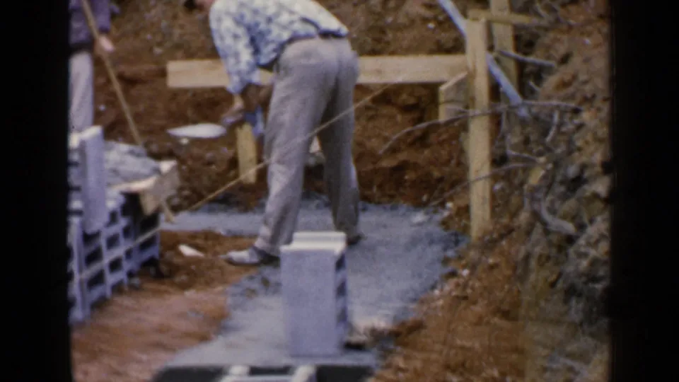 In 1957 Birmingham, Alabama footage, a man bends over a foundation area beside concrete blocks, wet cement, exposed dirt, and wooden supports.