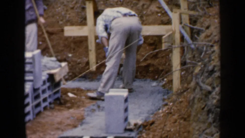 The worker continues shaping or spreading cement on a small foundation area in the Birmingham, Alabama home-movie clip.