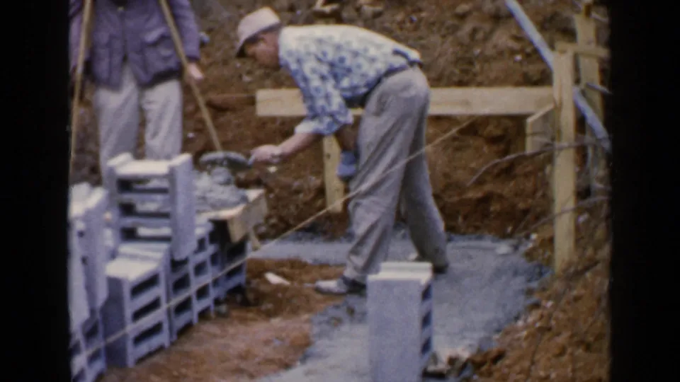 A man in a cap uses a trowel near stacked blocks, with another adult partly visible at the edge of the 1957 construction scene.