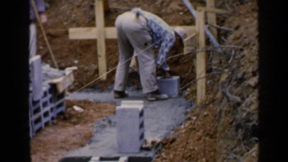 The worker bends beside a bucket and wet cement, surrounded by blocks, dirt, brush, and simple wooden supports in the Birmingham footage.