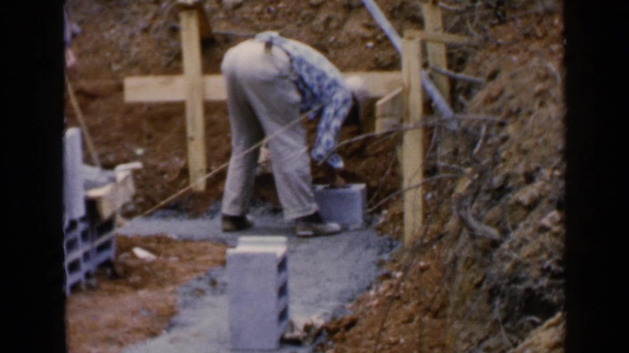 Worker bends over cinder blocks beside a concrete footing at an outdoor construction site.