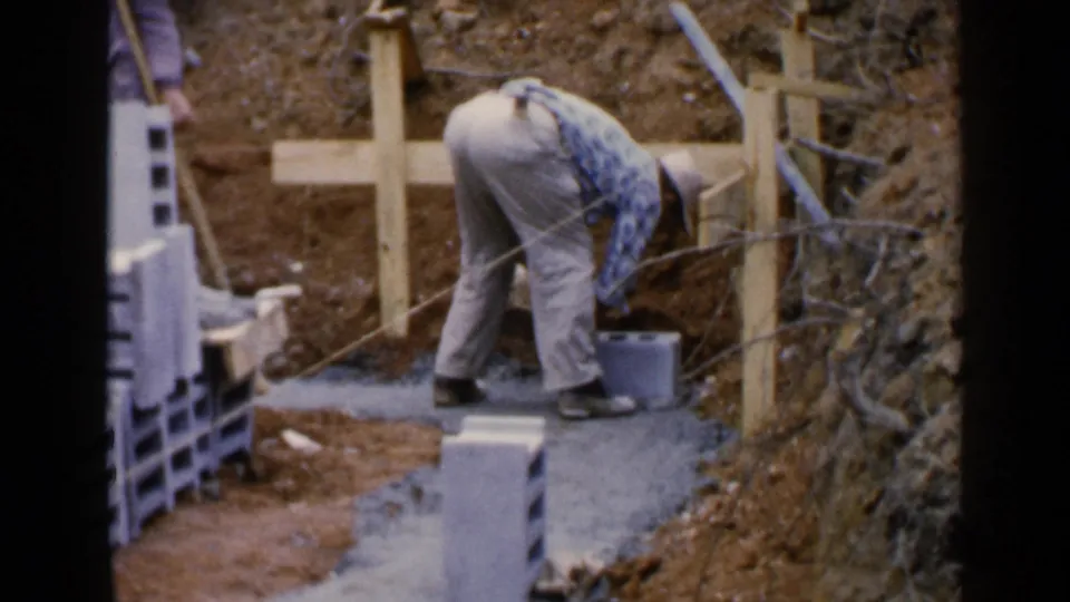 The frame shows the worker bent over in the construction trench, with stacked blocks, wooden form boards, and exposed soil visible around the footing.