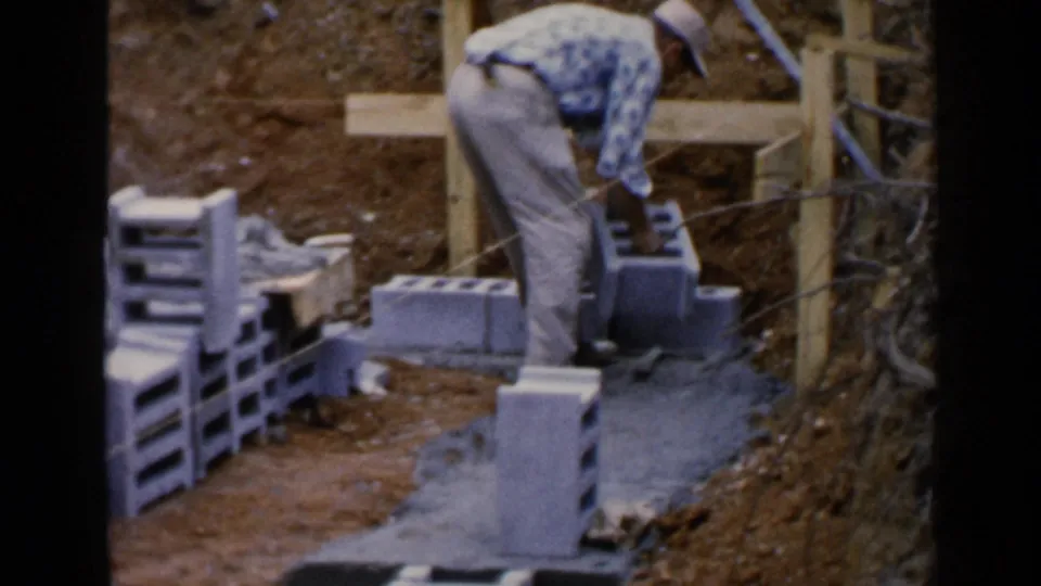 At the end of the clip, the worker stands over a cinder block near the concrete foundation, with stacks of blocks and wood formwork nearby.