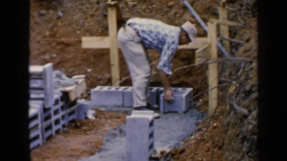 A man works along a row of grey blocks at a Birmingham construction site, with wood supports and a dirt slope behind him.