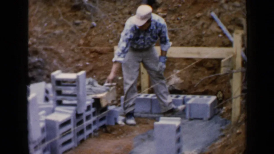 The worker stands over grey blocks in 1957 Birmingham footage, with a dirt bank, wood framing, and construction materials around him.