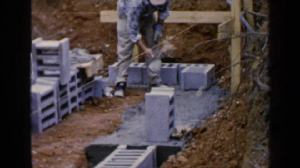 The 1957 home-movie frame shows a man positioning blocks along a string line at a dirt construction site in Birmingham.