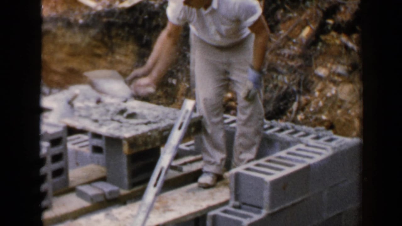 In 1957 Birmingham, Alabama footage, a man in a white shirt and khaki pants works with cinder blocks beside planks and exposed soil.