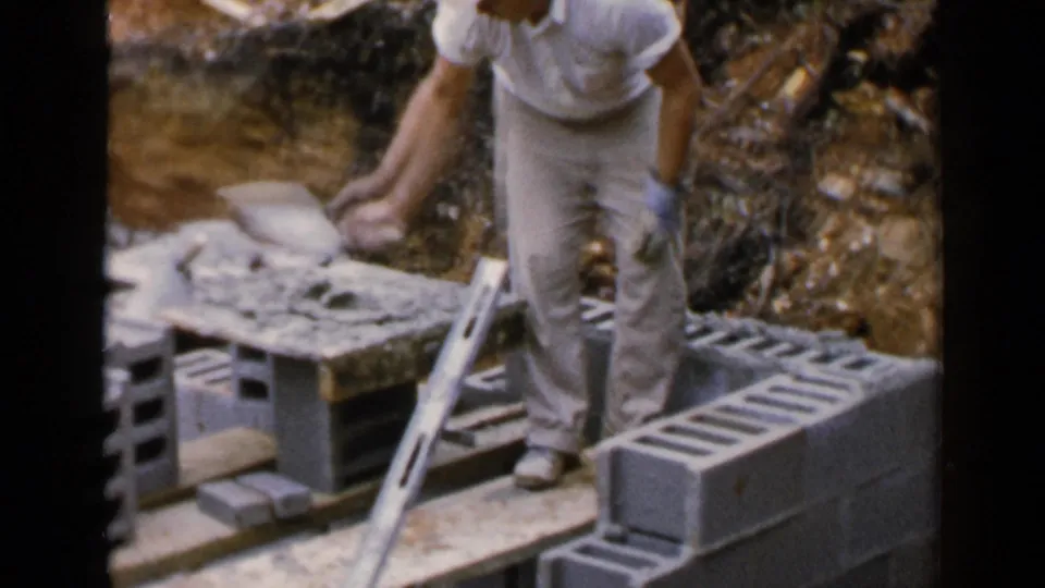 In 1957 Birmingham, Alabama footage, a man in a white shirt and khaki pants works with cinder blocks beside planks and exposed soil.