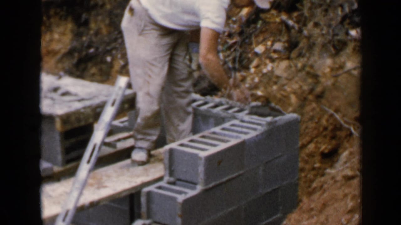 Man bends over a cinder block wall with a trowel near wooden planks and earth.