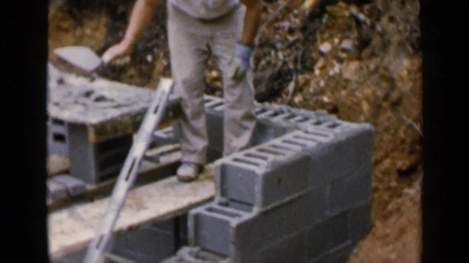 The 1957 home-movie frame shows a man lifting or setting a cinder block beside a low wall, with a level and planks nearby.