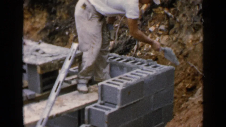 A man in khaki pants reaches across a cinder block wall with a trowel during a small construction project in Birmingham, Alabama.