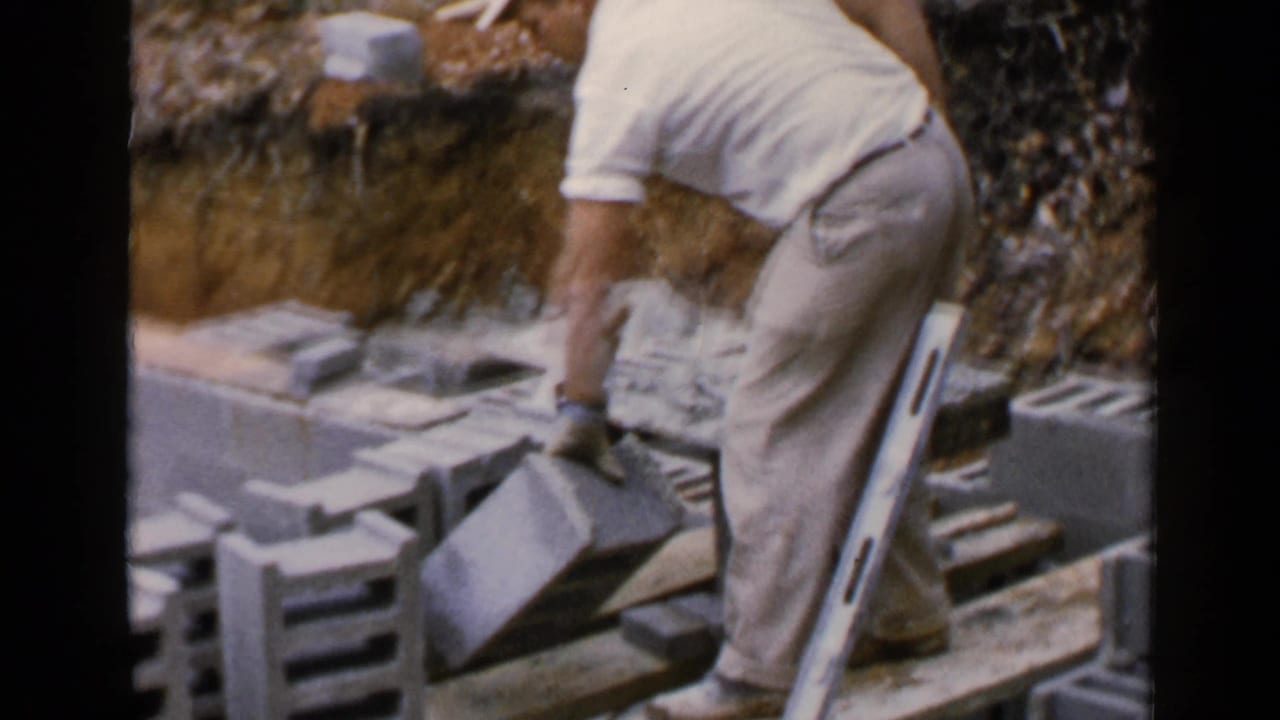 In 1957 Birmingham footage, a man in white work clothes bends over concrete blocks while applying mortar near a leaning level.