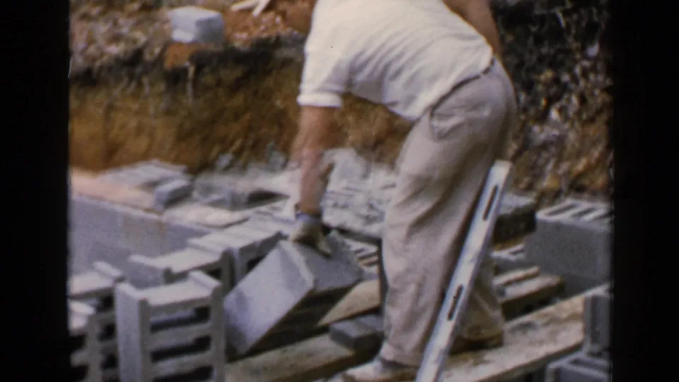 In 1957 Birmingham footage, a man in white work clothes bends over concrete blocks while applying mortar near a leaning level.
