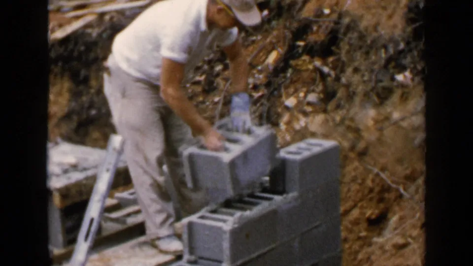 The worker places or adjusts a concrete block on a partially built foundation wall at the Birmingham outdoor site.