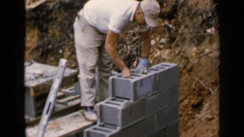 A man in light work clothes leans over stacked concrete blocks, continuing masonry work in the 1957 Birmingham home-movie scene.