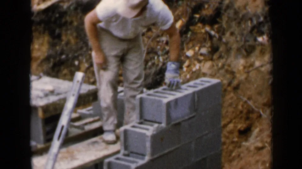 The worker stands over the concrete block foundation wall, with exposed soil and a leaning level visible in the wooded construction area.