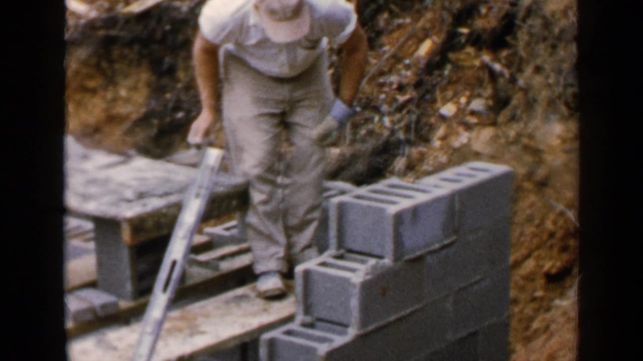Worker stands beside a partially built cinder block wall with a trowel nearby.