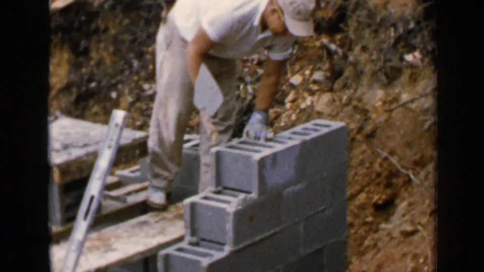 The 1957 home-movie frame shows the worker leaning over the wall as he continues the masonry process.