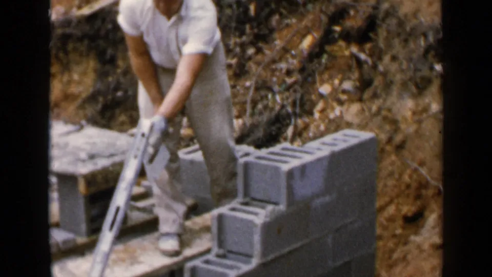 The worker stands with a trowel beside the partially built wall in the Birmingham 1957 construction footage.