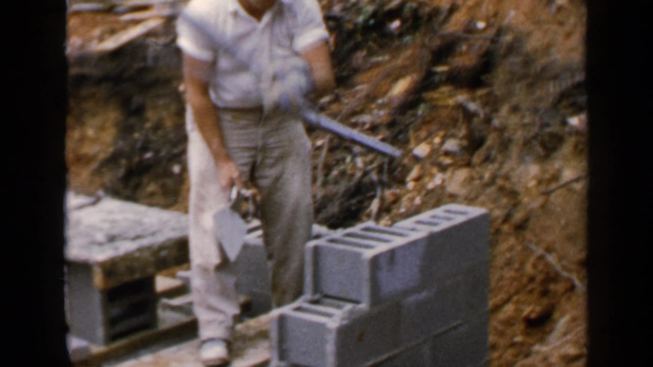 Worker stands beside a concrete block wall holding masonry tools at a construction site.