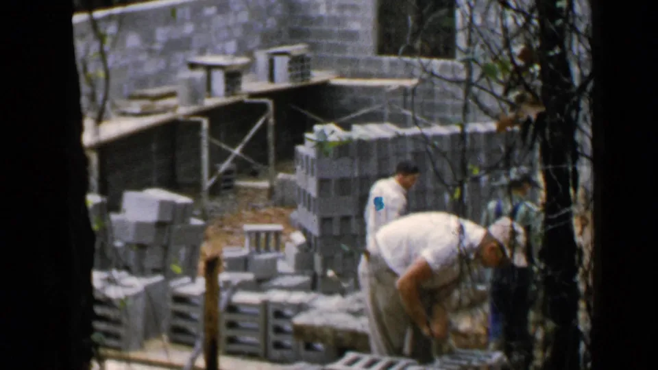 Wider 1957 Birmingham home-movie view of workers among stacked blocks, scaffolding, and unfinished masonry.