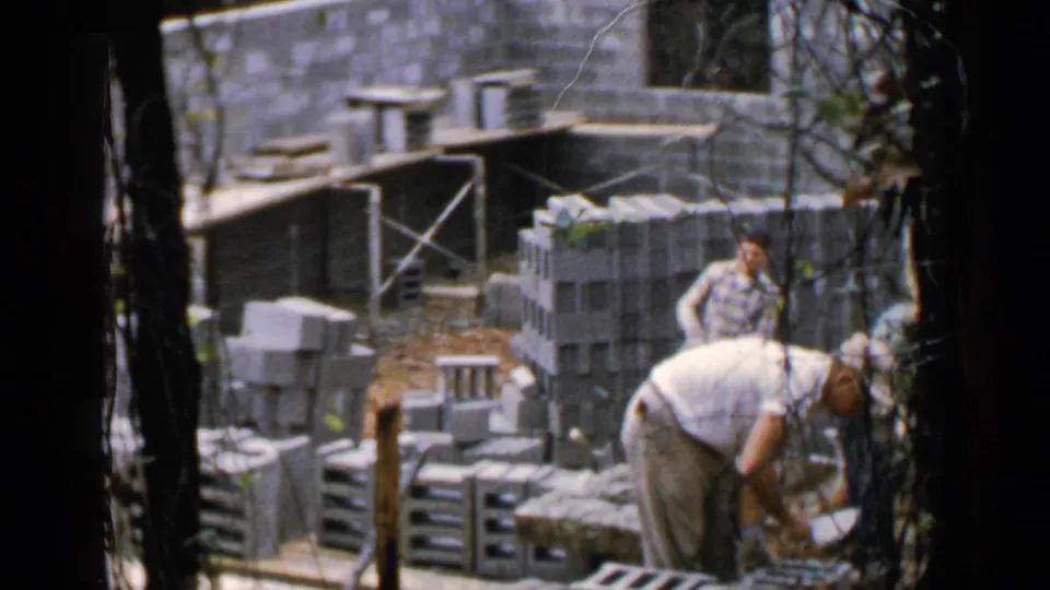 1957 Birmingham construction scene with workers near stacked cinder blocks, timber supports, and a partially built masonry wall.