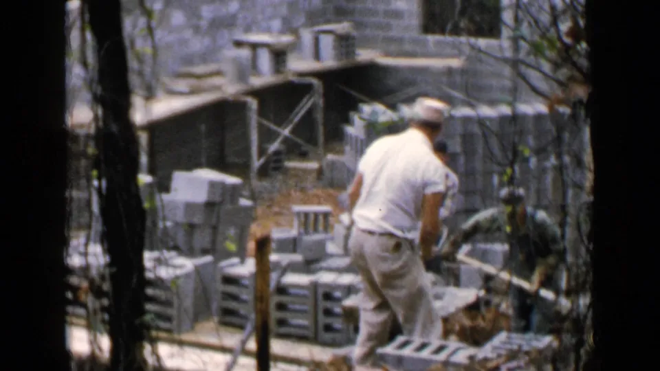 Worker in light clothing moves a wheelbarrow through a Birmingham cinder block construction site in 1957 footage.