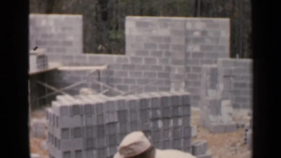 A worker in a light-colored cap is partly visible at the edge of a Birmingham construction frame with stacked blocks and unfinished masonry walls.