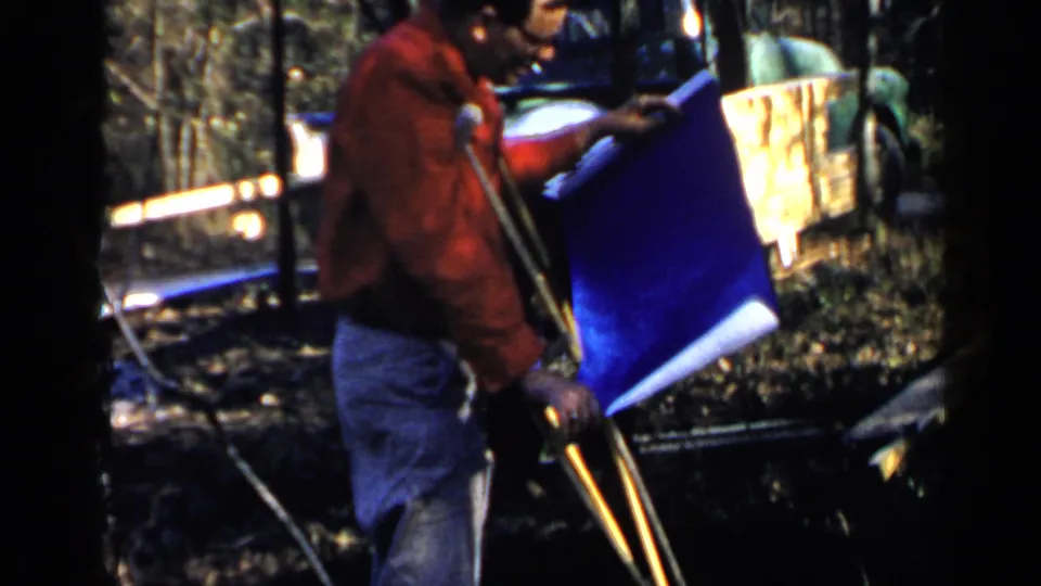 1957 Birmingham frame showing a man in a red jacket with crutches holding a blue plan sheet near another man and construction materials.