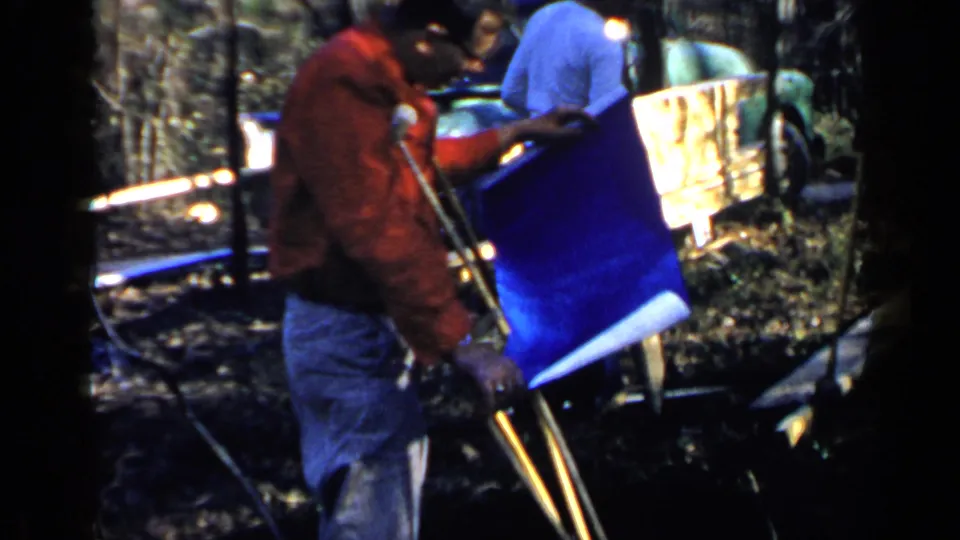 1957 Birmingham home-movie frame of two men beside a blue plan sheet, crutches, work supports, and a partially built backyard wall.