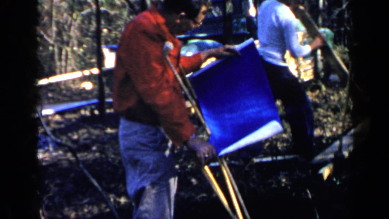 Man in a red jacket using crutches beside a large blue plan sheet in a wooded area.