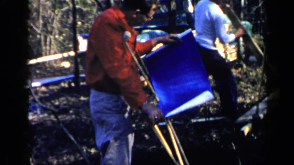 1957 Birmingham home-movie frame of a man on crutches looking down near a large blue plan sheet outdoors, with trees and equipment behind him.