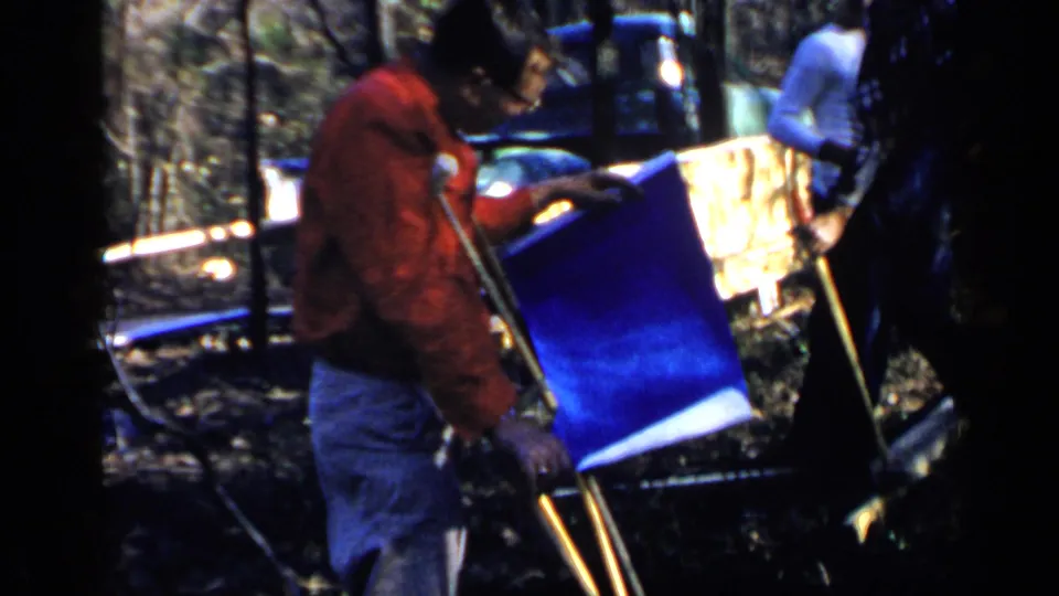 1957 Birmingham outdoor frame with a man on crutches examining a blue plan sheet while an adult in a white shirt stands near trees and gear.