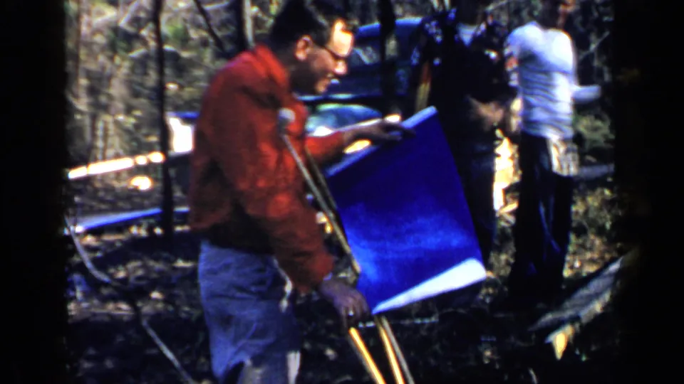A 1957 Birmingham 8mm frame shows a man in a red jacket using crutches and studying a blue plan sheet, with another adult and trees behind him.