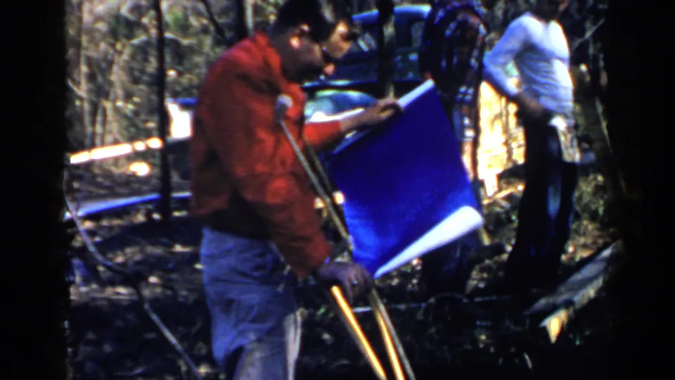 1957 Birmingham frame of a man using crutches beside a blue plan sheet, with an adult in a white shirt, trees, and outdoor materials nearby.