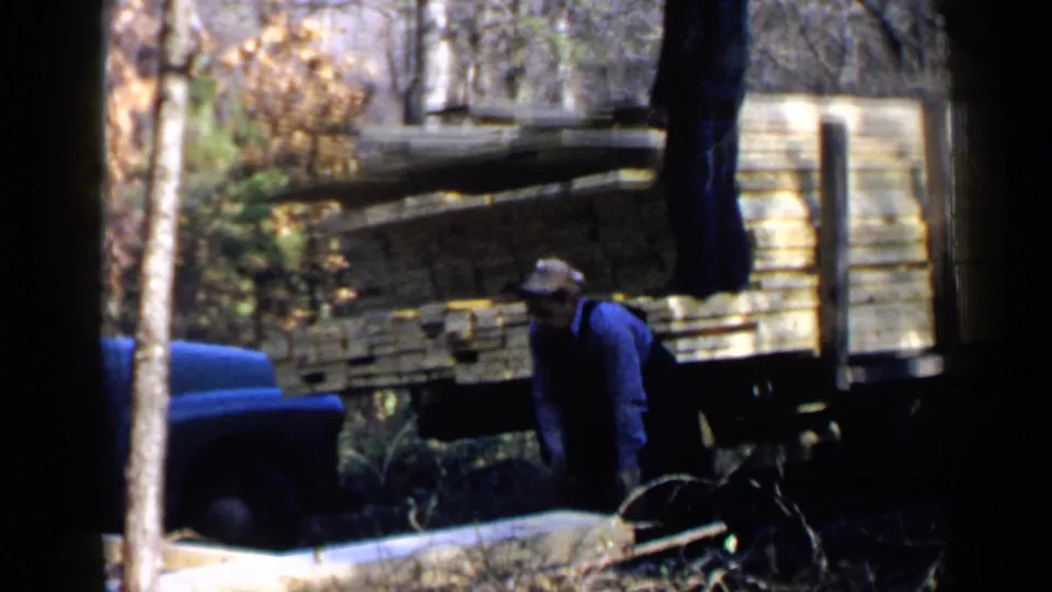 A capped worker stands beside lumber and a wooden truck bed, with a blue vehicle partly visible in the 1957 Birmingham footage.