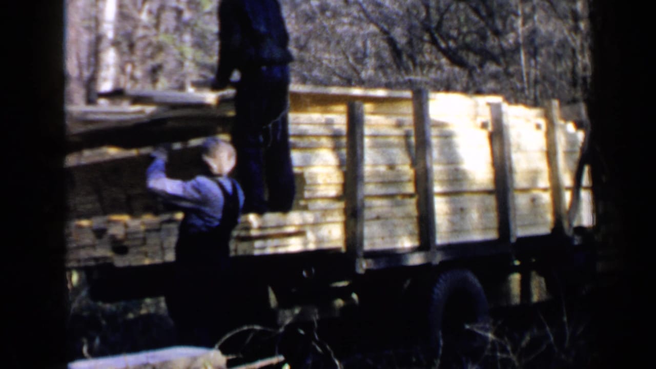 Two men unload lumber from a flatbed truck in a wooded area.