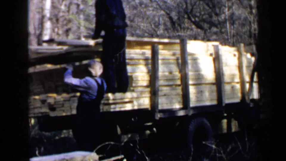 1957 Birmingham home-movie frame showing two men handling lumber beside a flatbed truck among bare trees.
