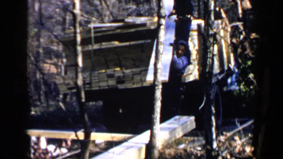 1957 Birmingham-area 8mm frame showing a man in a blue shirt near stacked or extended lumber beside a truck among trees.